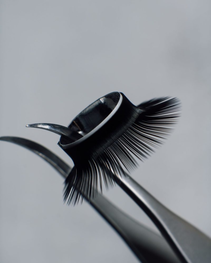 Close up of tweezers with ring of black artificial eyelashes hanging on tip, against gray background. Tool for lash extension that separating cilia and assisting create glamour look. Vertical.
